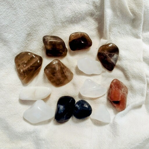 About a dozen partially tumbled rocks on a white cloth. About half are smokey quartz, some are white quartz, two are blueish gneiss and one is reddish gneiss.