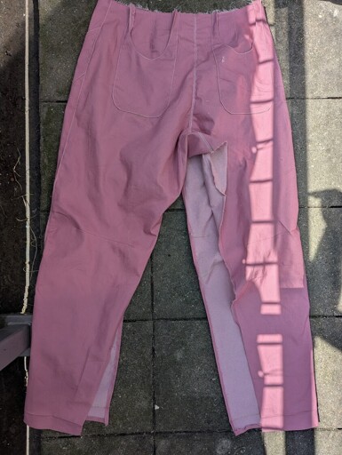 A pair of pink climbing pants laid flat on a stone patio in bright sunlight, showing the curved front pocket shapes and center front seams marked with light-colored thread. Strong shadows from a balcony railing fall across the fabric.