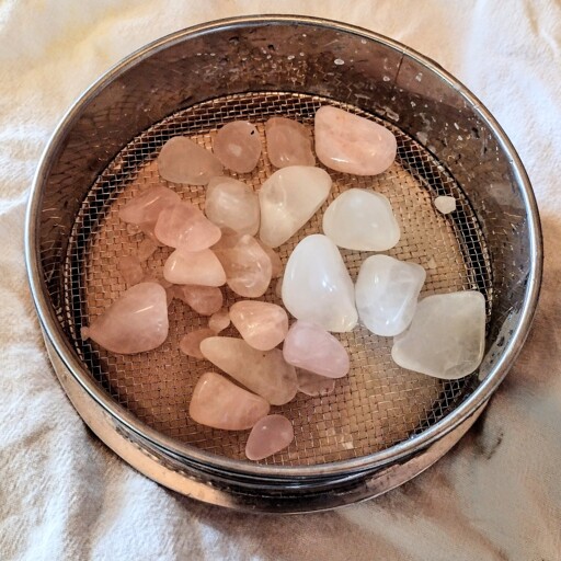 Wet, pink rose quartz and semi-white quartz tumbled stones in a metal sieve on top of a white cloth.