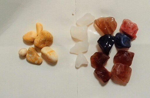 Rocks in three piles on a piece of white paper. The left are beige, unknown, the middle are white quartz, and the right are rougher smokey quartz with a couple darker and one reddish unknown rock.