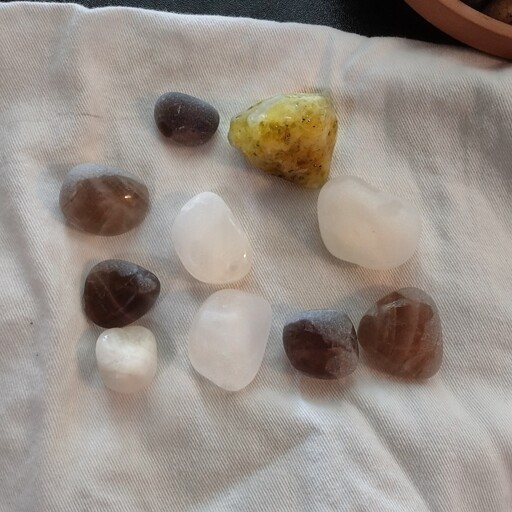 A collection of roughly tumbled rocks on a white napkin. Four are white quartz, five are smokey quarts, one is rougher and green of unknown type.